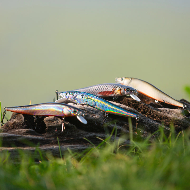Cargue la imagen en el visor de la galería, Four fishing lures designed to look like small fish, displayed on a piece of driftwood outdoors with green grass in the foreground and a blurred background.
