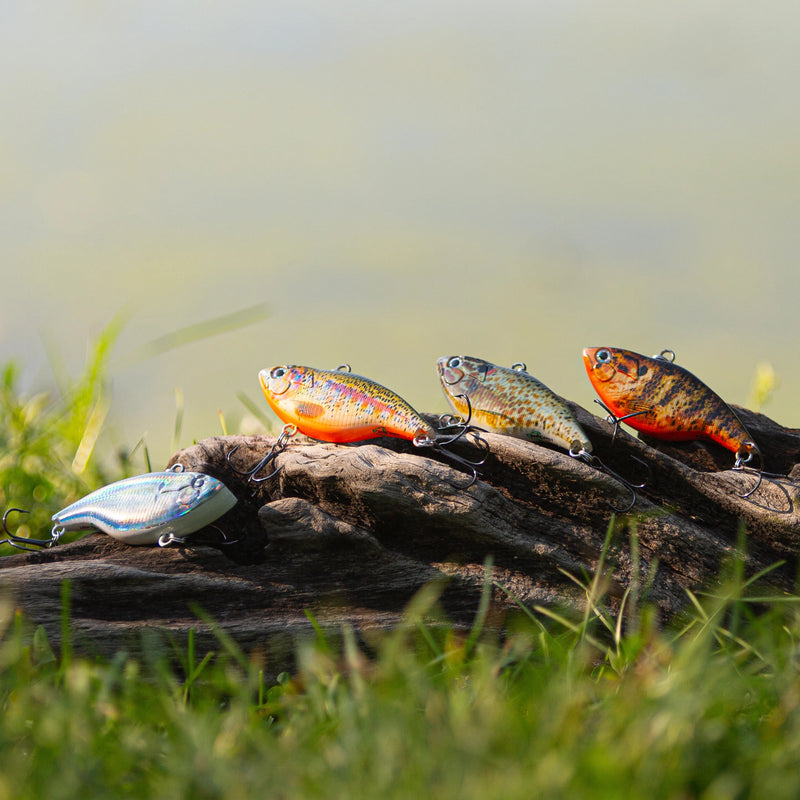 Cargue la imagen en el visor de la galería, Four live fishing lures shaped like small fish, including two orange-red ones, two silver-gray ones, and one blue one, resting on a piece of driftwood in a grassy outdoor setting.