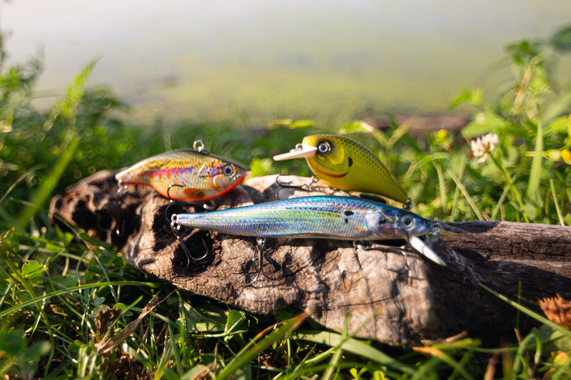 Load image into Gallery viewer, Three fishing lures displayed on a log outdoors in grassy water, including a silver-blue baitfish, a yellow topwater lure, and a multicolor crankbait.
