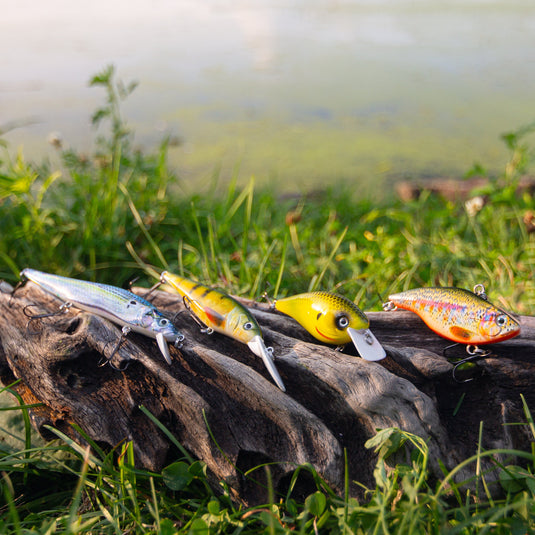 Three fishing lures in silver, yellow, and orange resting on a piece of driftwood in a grassy lakeside setting.
